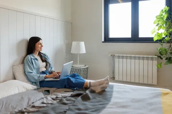 A smiling young woman sits comfortably on a bed with a laptop on her knees, working or browsing.